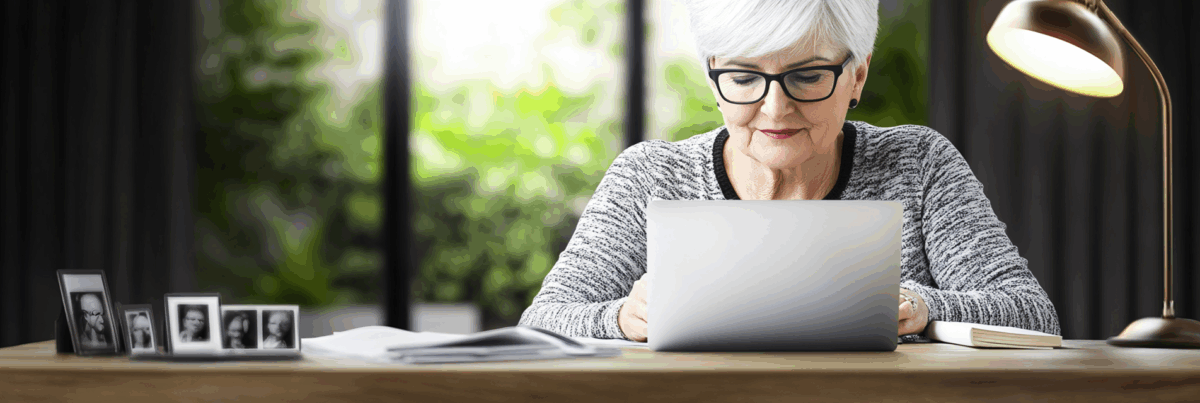 A woman in her 60s writing at a desk, lit by soft evening light, surrounded by notebooks and personal items, evoking reflection and nostalgia.