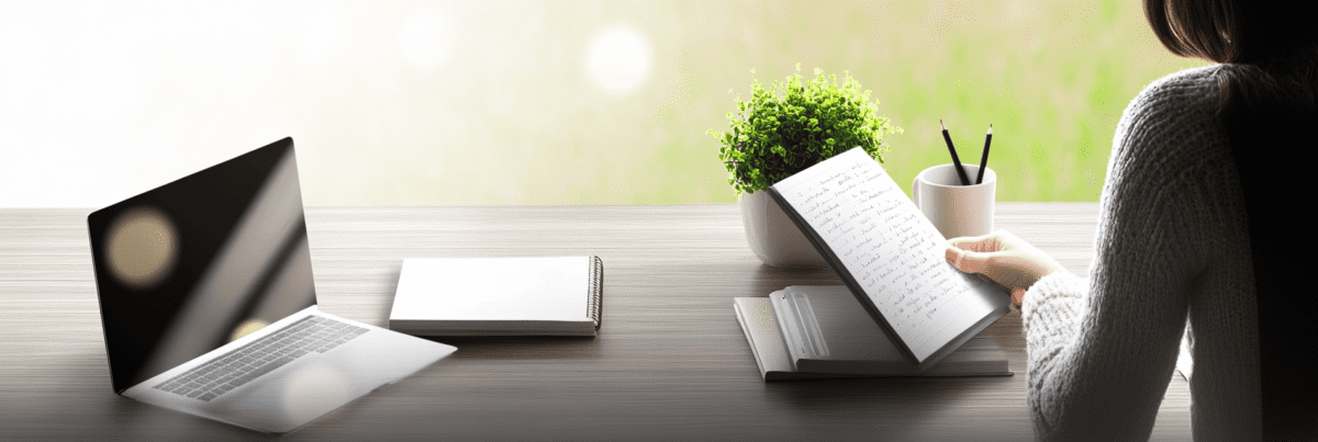 Over-the-shoulder view of a woman holding a handwritten notebook at a clean desk with a laptop and a plant, suggesting a quiet moment of reflection and revisiting creative ideas.