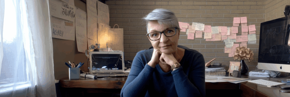 A thoughtful woman with short gray hair and glasses sitting at a creative desk space, surrounded by sticky notes, a laptop, and a desktop computer, with soft natural light filtering through a curtain.