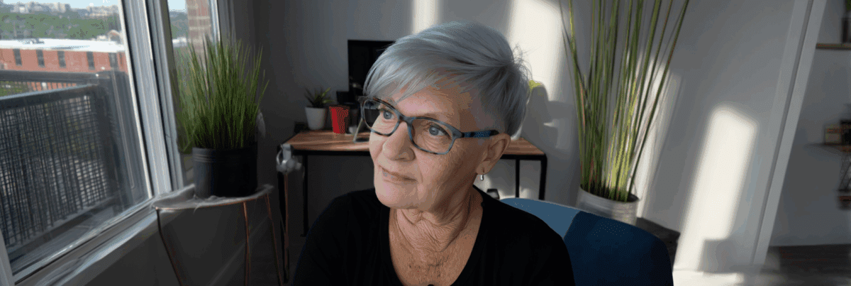 A woman with short silver hair and glasses sits in a sunlit room, gazing out the window with a thoughtful expression. The space around her is calm and personal, with plants, books, and soft shadows suggesting quiet reflection.