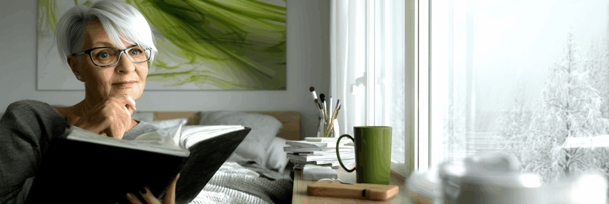 A woman with short white hair and glasses sits by a window, journaling thoughtfully with a soft winter landscape outside.