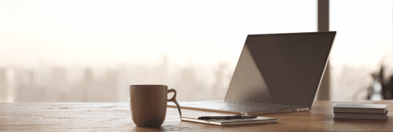 A minimalist workspace with a ceramic mug, closed notebook, pen, and sleek laptop on a wooden desk. The soft glow of early morning light filters through a window, blurring the city skyline in the distance. Calm and focused atmosphere.