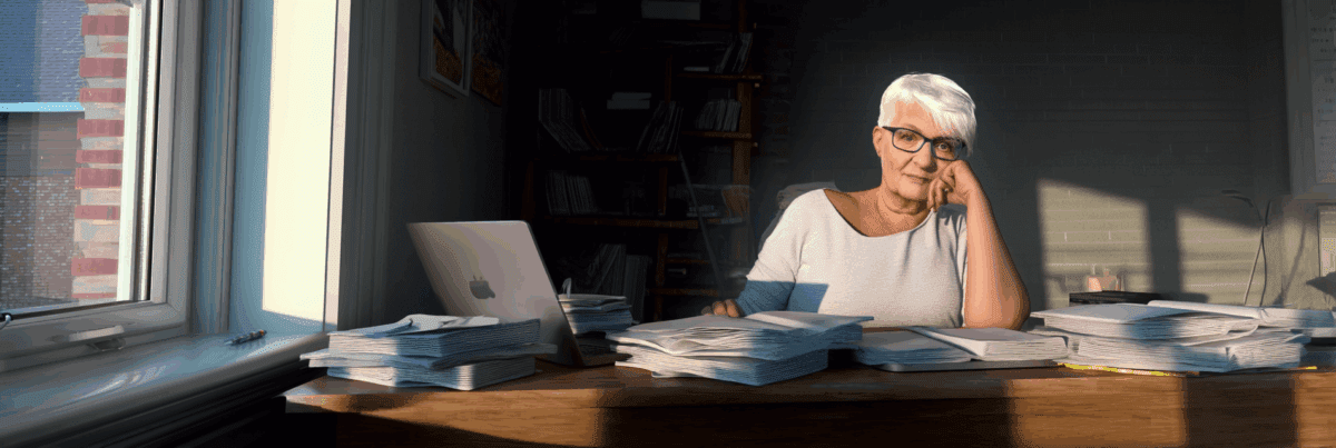 A white-haired woman with glasses sits at a desk covered in stacks of paper, resting her face on her hand as she gazes forward. Warm light from a window casts long shadows across the table, suggesting quiet reflection and creative tension.