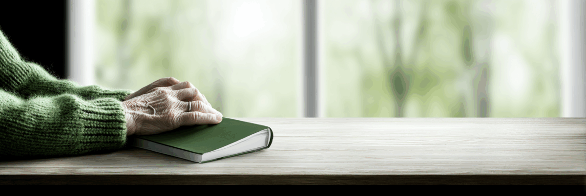 A close-up of an older woman’s hand resting gently on a closed green notebook, placed on a light wooden table in front of a softly blurred window. The scene is calm, reflective, and bathed in natural light.