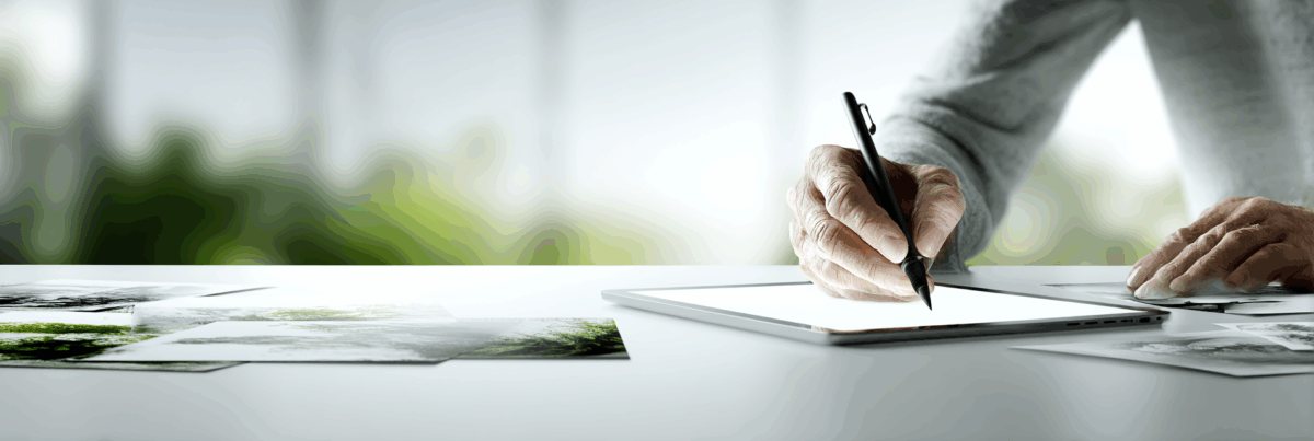 Close-up of hands holding a digital stylus and working on a tablet, with scattered printed photos on the table. The background is softly blurred greenery, suggesting a calm and creative workspace.