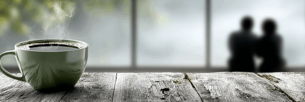 A steaming green coffee cup sits on a rustic wooden table in sharp focus, while two blurred figures sit close together in the background, creating an atmosphere of warmth, trust, and quiet connection.