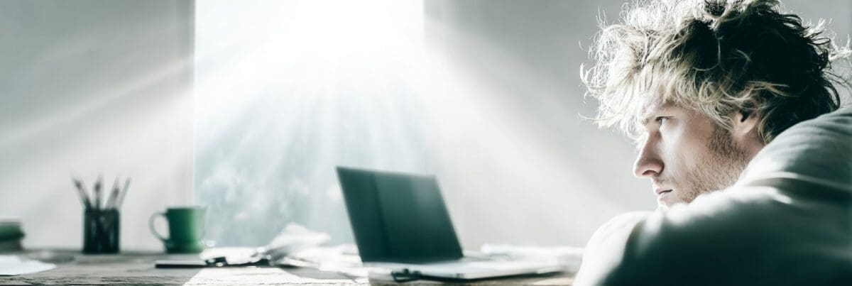 A person sits at a sunlit desk, gazing thoughtfully toward a laptop as bright morning light streams through the window. A green mug, papers, and pencils rest nearby, creating a quiet, reflective atmosphere.
