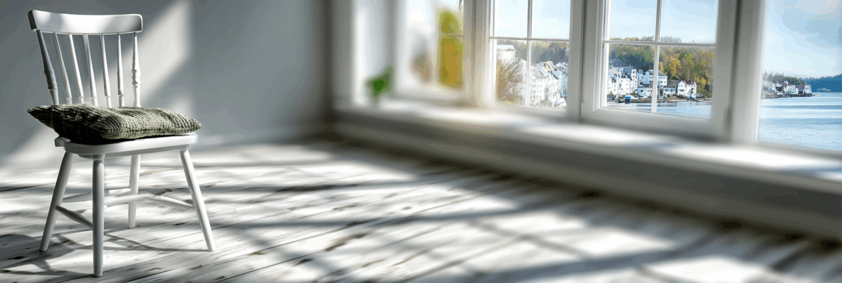 A white wooden chair with a green knitted cushion sits in soft sunlight near a wide window. Light spills across the worn wooden floor, and a quiet town and water are visible outside.