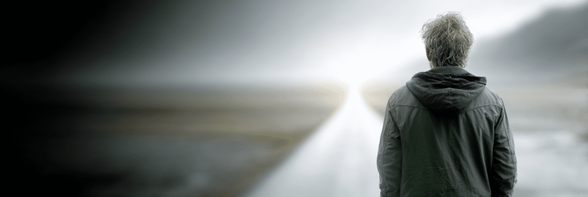 A person seen from behind standing on a pale road that stretches forward into a softly blurred landscape, with muted light and distant hills in the background.