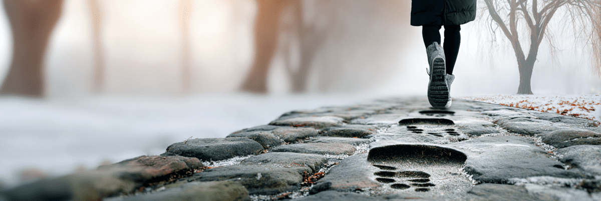 A person walking away along a cobblestone path in winter, visible footprints continuing forward on the stone surface, bare trees and soft fog in the background.