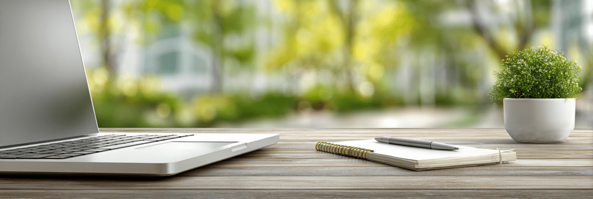 Open laptop on a wooden desk beside a notebook and pen, with soft natural light and a blurred green outdoor background, suggesting a quiet space for thinking and writing