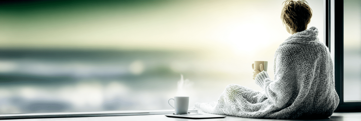 A person wrapped in a blanket sitting quietly by a large window holding a cup of coffee, reflecting in silence after digital overwhelm and constant online noise.