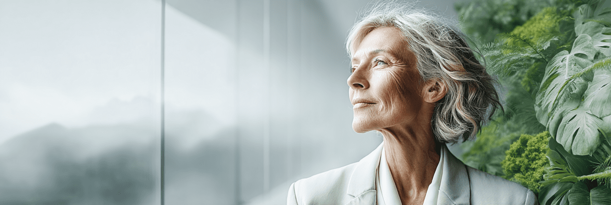 A thoughtful older woman stands beside a large window, looking toward the light. Behind her, lush green foliage contrasts with the clean modern interior, creating a sense of reflection, growth, and looking toward the future.