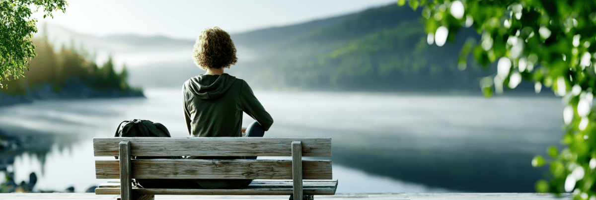 A person sits alone on a wooden bench overlooking a calm lake at sunrise, with a backpack beside them, pausing in quiet contemplation before continuing their journey.