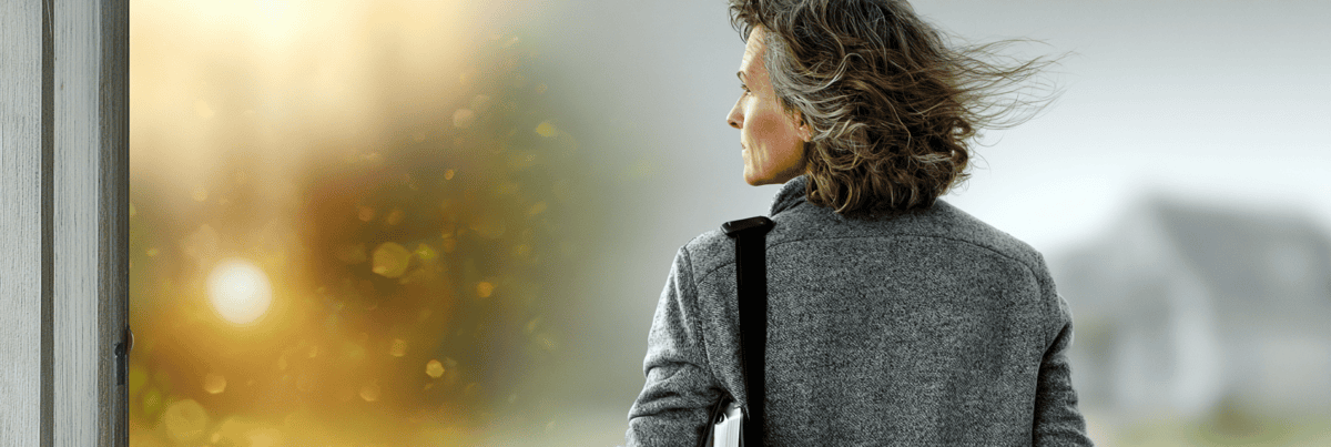 A mature woman stands outdoors in the morning light, looking thoughtfully toward the horizon with a notebook tucked under her arm.